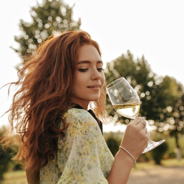 Tender girl with ginger long hair and lovely freckles in fashionable summer green clothes looking d Tender girl with ginger long hair and lovely freckles in fashionable summer green clothes looking down and holding glass with wine outdoor..