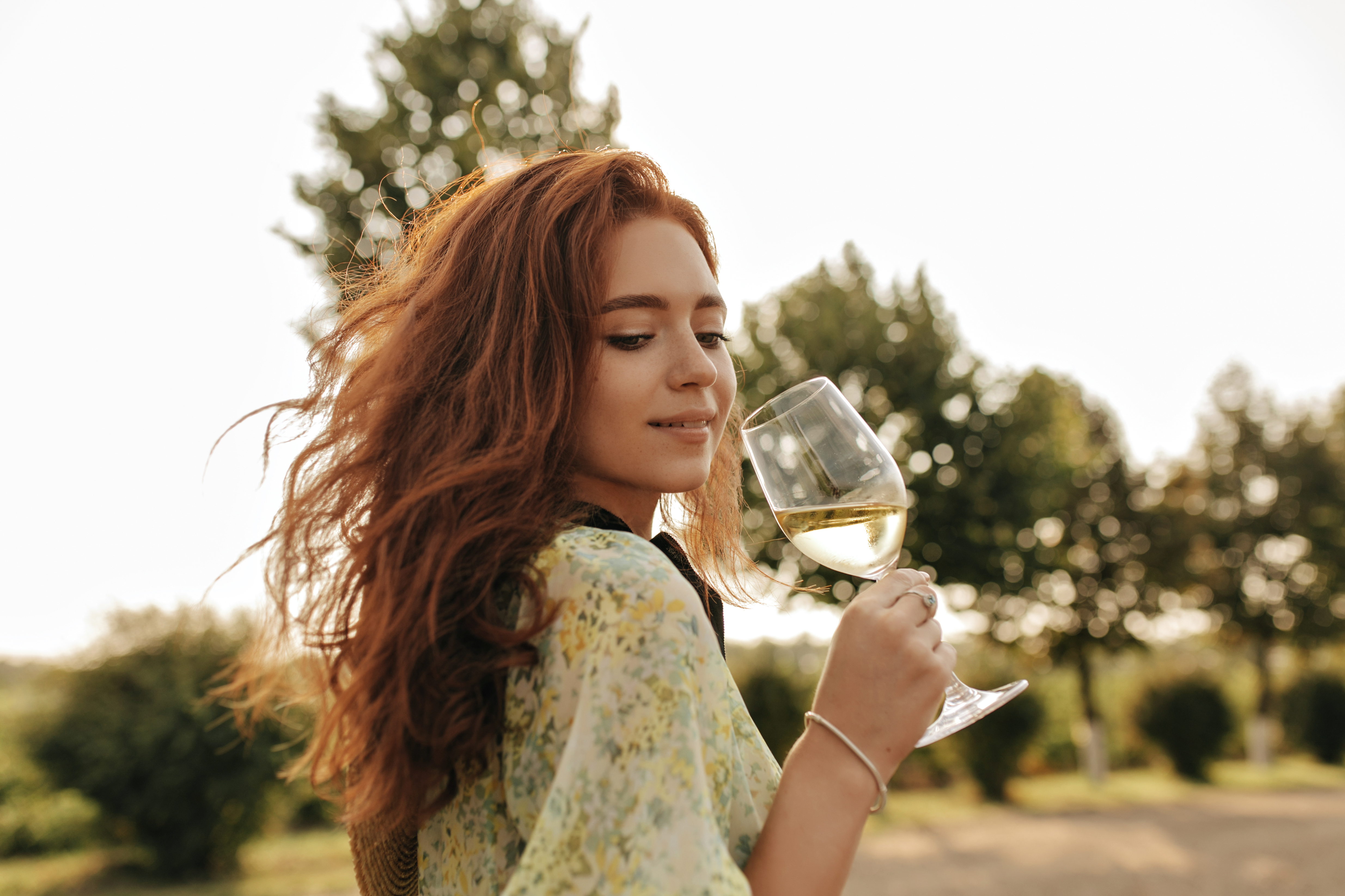Tender girl with ginger long hair and lovely freckles in fashionable summer green clothes looking down and holding glass with wine outdoor..